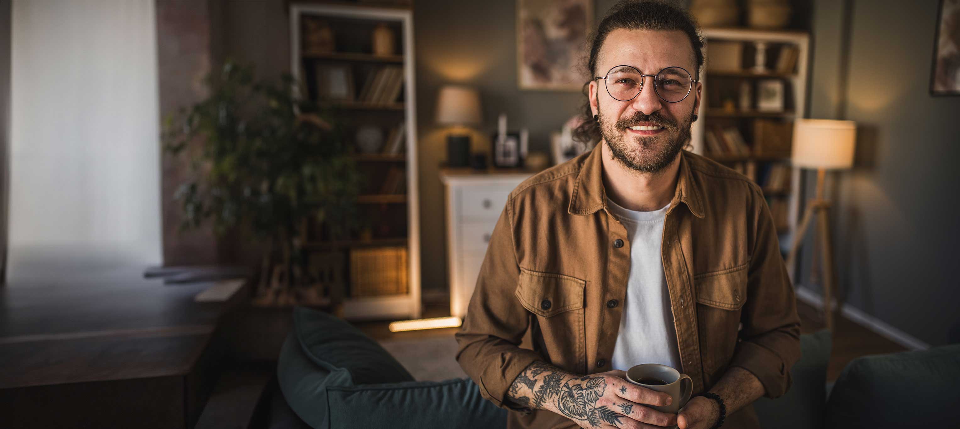 Foto - Ein tätowierter Mann mit einer Brille hält einen Kaffee in beiden Händen und sitzt in seinem Wohnzimmer.